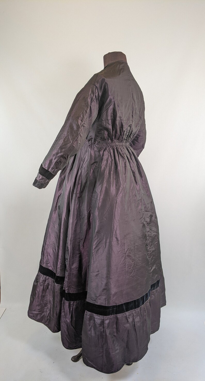 May include: A long, dark purple dress with a high collar and long sleeves. The dress features a gathered waist and a full skirt with black velvet trim at the hem. The fabric has a subtle sheen.