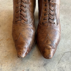 K&ouml;nnte beinhalten: Ein Paar braune Lederstiefel mit Schn&uuml;rung. Die Stiefel haben eine spitze Zehenpartie und ein abgenutztes Aussehen.