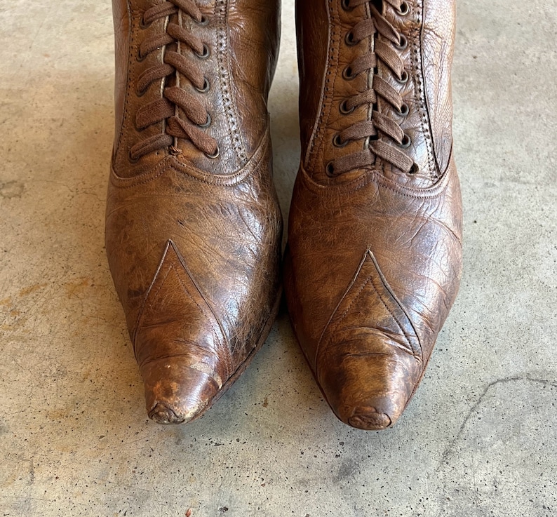 K&ouml;nnte beinhalten: Ein Paar braune Lederstiefel mit Schn&uuml;rung. Die Stiefel haben eine spitze Zehenpartie und ein abgenutztes Aussehen.