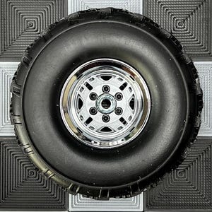May include: A black rubber tyre with a chrome rim, featuring a six-bolt pattern, rests on a black and white chequered surface. The tyre has a rugged tread design. The rim is shiny and reflects light. The tyre is suitable for off-road use.