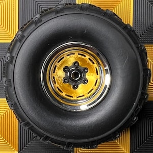 May include: A black rubber tire with a gold-colored rim and a silver-colored outer ring. The tire is set against a background of alternating gold and black square tiles.