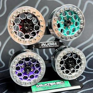 May include: Four 1.9" beadlock wheels in a display stand. The wheels have silver rims and various colored centers: red, teal, purple, and black. The display stand has the text "INJORA" and "MILKTEARC CUSTOM SCALE PRODUCTS".
