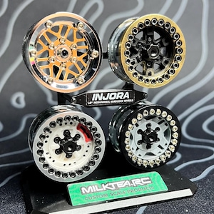 May include: Four 1.9" beadlock wheels displayed on a black stand. The wheels have various designs and colors, including gold, black, silver, and white. The stand has the "INJORA" logo and the "MILKTEA.RC" logo.