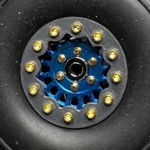 May include: Close-up of a black rubber tire with a blue and gold metal wheel. The wheel has a circular design with gold bolts around the perimeter. The tire has a deep tread pattern.