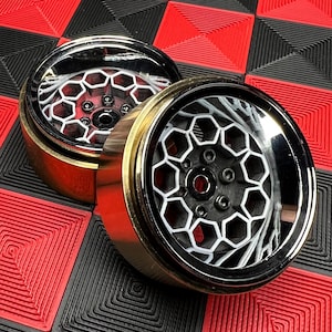 Può includere: Due ruote di auto in miniatura cromate e dorate con un design a nido d'ape bianco. Le ruote hanno un centro nero e sono poste su uno sfondo a scacchi rosso e nero.