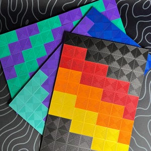 Op de afbeelding: Een verzameling van drie rechthoekige panelen met een geometrisch raster van verhoogde, piramidevormige vierkanten. Eén paneel toont een verloop van rood, oranje, geel en zwart, terwijl de andere twee panelen tinten paars, blauw en groenblauw bevatten.