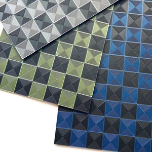 Op de afbeelding: Drie getextureerde vierkante matten in grijstinten, groen en blauw. Elke mat heeft een herhalend patroon van concentrische vierkanten, waardoor een 3D-effect ontstaat. De matten zijn in verschillende hoeken gerangschikt en tonen de kleurvariaties en het ontwerp.