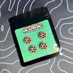 May include: Four small, brown custom wheel faces are displayed on a green card inside a black plastic bag. The card has the text "MILKTEA.RC" and "0.7" CUSTOM WHEEL FACE". The wheel faces have a circular design with multiple holes.