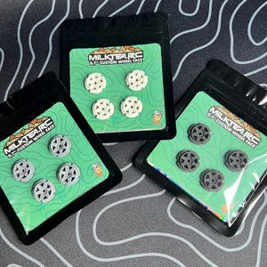 Op de afbeelding: Drie kleine zwarte plastic zakjes met elk vier 0,7" custom wielvlakken voor RC-auto's. De wielvlakken zijn wit, grijs en donkergrijs, met een stervormig ontwerp. Elke zak heeft een groene kaart met het "MILKTEA.RC" logo.