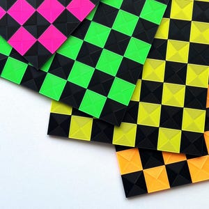 Op de afbeelding: Een verzameling kleurrijke, geometrisch gevormde vierkanten in neonroze, groen, geel en oranje, tegen een zwarte achtergrond. De vierkanten zijn gerangschikt in een dambordpatroon, met een getextureerd, driedimensionaal uiterlijk. De vierkanten liggen op een witte ondergrond.