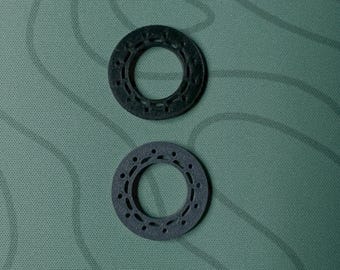 May include: Two dark gray, circular objects with a central hole and evenly spaced smaller holes around the perimeter. The objects are on a textured, green background.