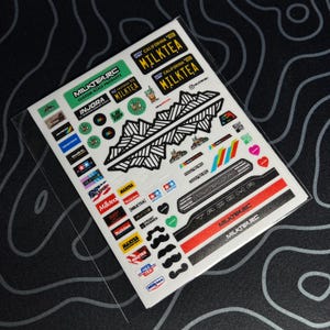 以下が含まれることがあります： さまざまなデザインとテキストが特徴の、カスタムスケール製品ステッカーのシートです。ステッカーには、「MILKTEA.RC」、「MILKTEA」、「CALIFORNIA」、「TACOMA」の文字が含まれています。ステッカーは透明なビニールスリーブに入っています。