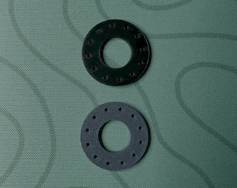 May include: Two dark gray, circular washers with central holes and evenly spaced smaller holes around the perimeter. One washer is above the other. The background is a textured, olive green surface.