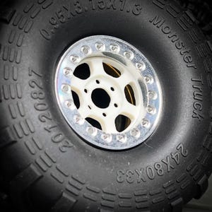 May include: Close-up of a black Monster Truck tire with a white and silver rim. The tire has deep treads and the text "Monster Truck" and "24X80X33" are visible. The rim has a series of small holes around its edge.