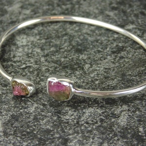 May include: A silver bangle bracelet with two pink and green gemstones set into the metal. The bracelet is open and has a simple design.