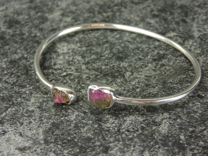 May include: A silver bangle bracelet with two pink and green gemstones set into the metal. The bracelet is open and has a simple design.