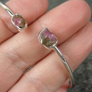 May include: A silver bangle bracelet with two raw, pink and green tourmaline gemstones set in the metal. The bracelet is open and has a simple design.