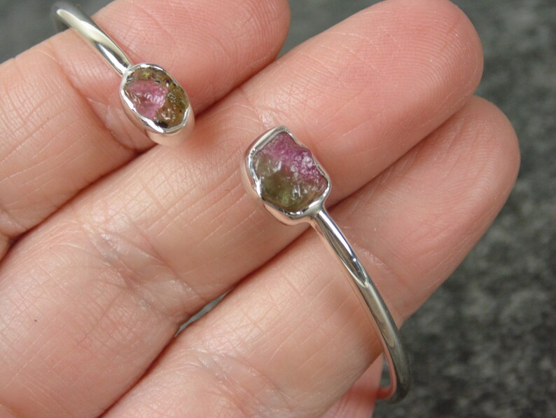 May include: A silver bangle bracelet with two raw, pink and green tourmaline gemstones set in the metal. The bracelet is open and has a simple design.