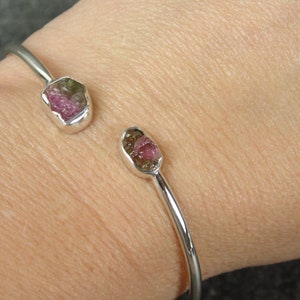 May include: A silver bangle bracelet with two pink and green tourmaline gemstones set in the metal. The bracelet is on a person's wrist.