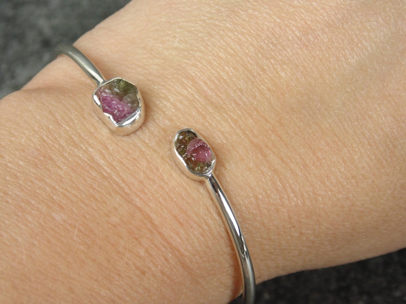 May include: A silver bangle bracelet with two pink and green tourmaline gemstones set in the metal. The bracelet is on a person's wrist.