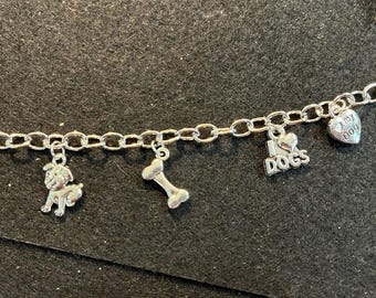 May include: A silver charm bracelet with dog-themed charms. The charms include a small dog figure, a bone, a heart with "I love dogs" text, and a heart-shaped charm with "my dog" text. The bracelet is displayed against a dark background.
