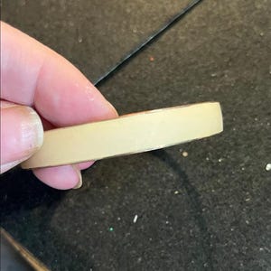 Op de afbeelding: Een beige, rechthoekig object met een metalen rand wordt tussen vingers gehouden. Het object lijkt een stuk tape of een soortgelijk kleefmateriaal te zijn. De achtergrond is een donker, mogelijk zwart oppervlak.