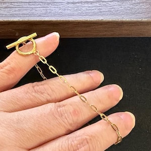 Op de afbeelding: Een goudkleurige ketting met een ovale schakelketting en een tuimelsluiting. De ketting wordt door een hand omhoog gehouden, wat het delicate ontwerp en de warme tint van het metaal laat zien. De sluiting is een cirkelvormige ring met een horizontale staaf.