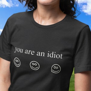 May include: Black t-shirt with the phrase "you are an idiot" in white text. Below the text are three smiling face emojis. The shirt is a crew neck style.