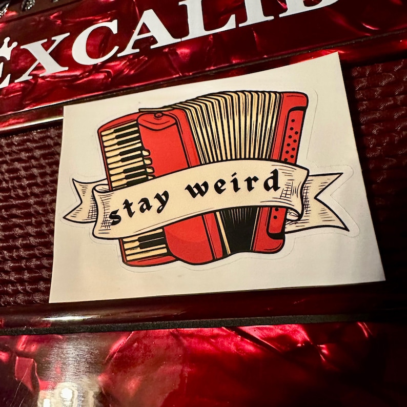 May include: A sticker featuring a red accordion with black and beige details, and the words "stay weird" on a banner. The sticker is placed on a red, textured surface.