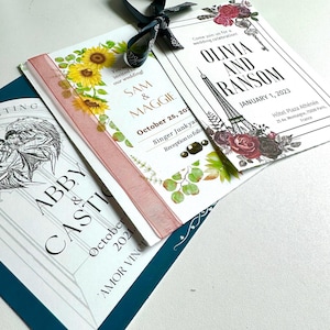 May include: Wedding invitation suite featuring three invitation cards. One card has sunflowers and the text "SAM & MAGGIE". Another card has the Eiffel Tower and the text "OLIVIA AND RANSOM". The third card has the text "ABBY & CASTIC". A pink ribbon and a dark green ribbon are attached.