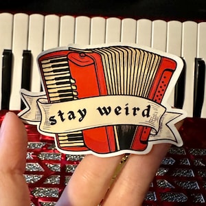 May include: A sticker featuring a red accordion with black and white keys, and the words "stay weird" on a banner. The sticker is held in front of a red and silver accordion.