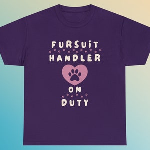 May include: A purple t-shirt with the text "FURSUIT HANDLER ON DUTY" in white. A pink heart with a paw print is centered below the word "HANDLER". Small paw prints surround the text.
