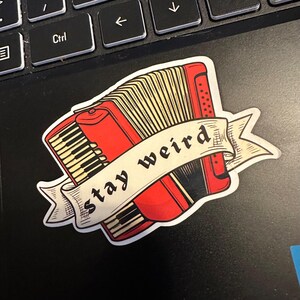 May include: A red accordion sticker with a banner that reads "stay weird." The sticker features a detailed illustration of an accordion with black and white keys and a beige bellows. The sticker is placed on a black surface.