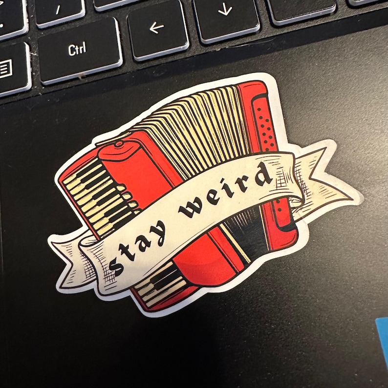 May include: A red accordion sticker with a banner that reads "stay weird." The sticker features a detailed illustration of an accordion with black and white keys and a beige bellows. The sticker is placed on a black surface.