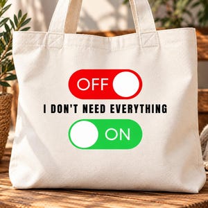 Puede incluir: Un bolso de tela color crema con un gráfico de dos interruptores. El interruptor rojo superior está en OFF, el verde inferior está en ON, y el texto I DON'T NEED EVERYTHING está impreso en negro entre ambos interruptores.