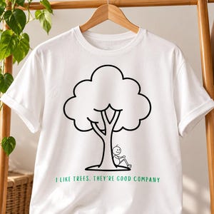 Puede incluir: Una camiseta blanca de manga corta con un dibujo de líneas negras de un árbol con una figura de palo sentada en su base. Debajo del gráfico, el texto en letras mayúsculas verdes dice: I LIKE TREES. THEY'RE GOOD COMPANY.