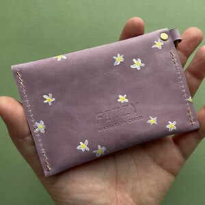 May include: A small, lavender-colored leather wallet with a white daisy pattern and the text "FITZY HANDMADE IN CANADA" embossed on the front.