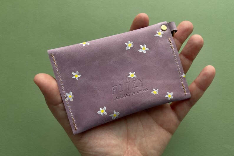 May include: A small, lavender-colored leather wallet with a white daisy pattern and the text "FITZY HANDMADE IN CANADA" embossed on the front.