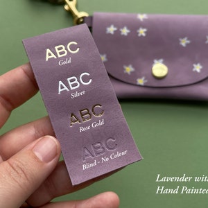May include: A close-up of a lavender leather wallet with hand-painted white flowers. The wallet has a flap closure and a gold button. A sample swatch of leather is shown with the text "ABC" in gold, silver, rose gold, and blind embossed lettering.
