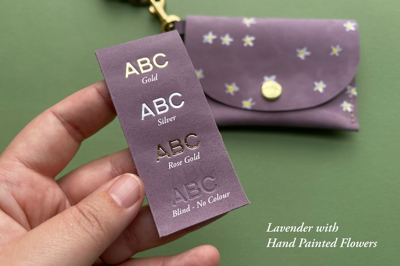 May include: A close-up of a lavender leather wallet with hand-painted white flowers. The wallet has a flap closure and a gold button. A sample swatch of leather is shown with the text "ABC" in gold, silver, rose gold, and blind embossed lettering.