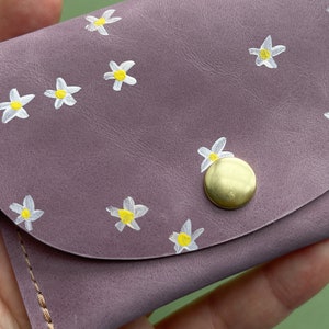May include: A small, purple leather wallet with a snap closure and a gold button. The wallet is decorated with a white daisy pattern.