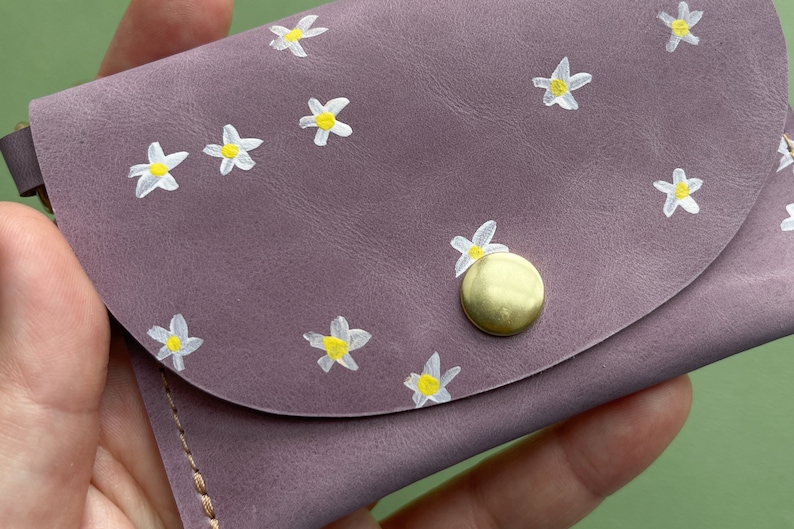 May include: A small, purple leather wallet with a snap closure and a gold button. The wallet is decorated with a white daisy pattern.
