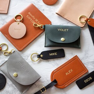 May include: Assortment of personalized leather goods, including keychains, wallets, and passport holders. The items are in various colors, including brown, black, green, and gray. The text on the items includes names, dates, and phrases like "home" and "boss".