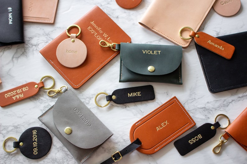 May include: Assortment of personalized leather goods, including keychains, wallets, and passport holders. The items are in various colors, including brown, black, green, and gray. The text on the items includes names, dates, and phrases like "home" and "boss".