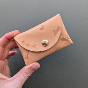 May include: A small, light tan leather wallet with a flap closure and a gold-colored snap. The wallet is embossed with heart and arrow designs. The wallet is held in a hand.