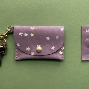 May include: A small, purple leather keychain wallet with a floral pattern and a gold key ring. The wallet has a flap with a gold button closure. The keychain has two keys attached to it. The wallet is on a green background.