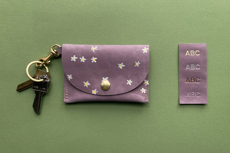 May include: A small, purple leather keychain wallet with a floral pattern and a gold key ring. The wallet has a flap with a gold button closure. The keychain has two keys attached to it. The wallet is on a green background.