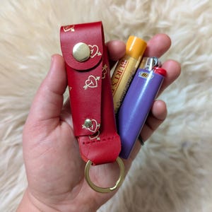 May include: A red leather keychain with a gold ring and a heart pattern. The keychain is holding a yellow tube of Burt's Bees lip balm and a purple lighter.