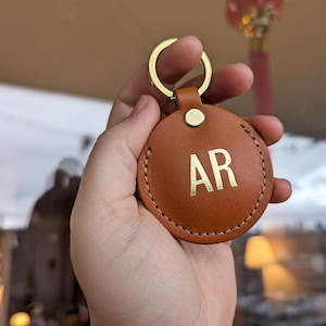 Puede incluir: Un llavero de cuero marrón con un anillo de metal dorado y una etiqueta redonda de cuero con las iniciales "AR" grabadas en oro.