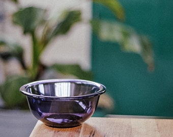 May include: A small, dark purple glass bowl with a rounded rim sits on a wooden surface. The bowl reflects the light, and the background includes a green wall and a blurred plant.